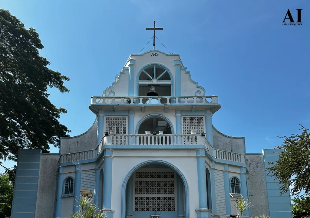 A detailed close-up of the upper facade of St. Nicholas of Bari Parish in Siaton, Negros Oriental, highlighting the central bell tower with its bronze bell, white balustrades, and intricate ventilation blocks under a clear sky.