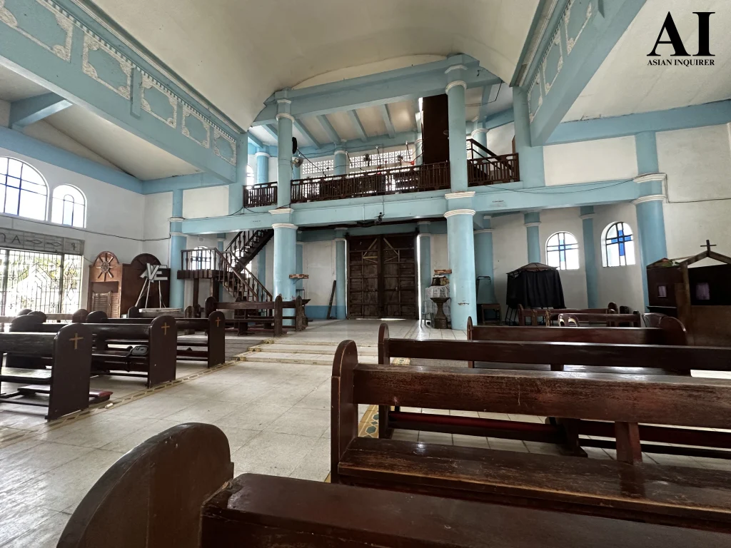A wide-angle interior view facing the entrance of St. Nicholas of Bari Parish, featuring a dark wood choir loft with a staircase, blue structural pillars, and heavy timber main doors.