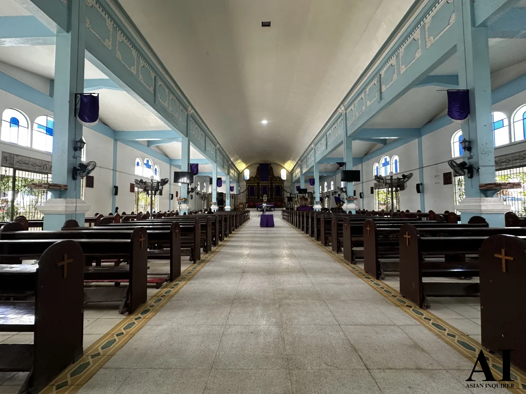 A wide-angle interior view of St. Nicholas of Bari Parish in Siaton, Negros Oriental, showing the long central aisle, rows of wooden pews, and the high barrel vault ceiling supported by sky-blue pillars leading to the altar.