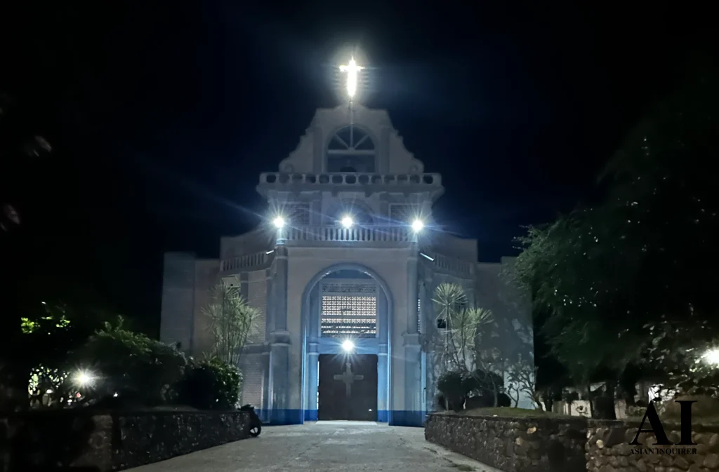 A nighttime view of the St. Nicholas of Bari Parish in Siaton, Negros Oriental, showing the facade illuminated by floodlights and a glowing white cross at the top of the bell tower against a dark sky.