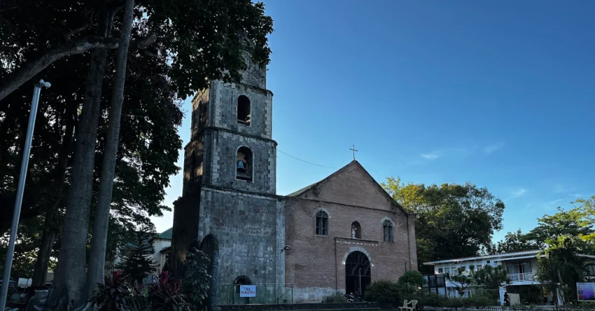 Bacong Church in Negros Oriental with a tall old stone bell tower, brick church facade, arched front entrance, cross on the roof, large trees on the left, and a clear blue sky above the quiet church grounds.