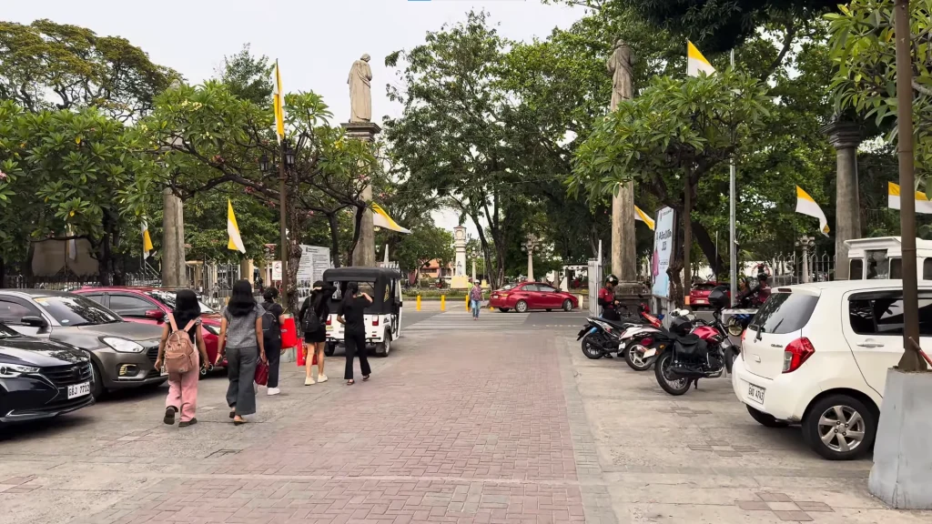 Street-level view of the entrance to the St. Catherine of Alexandria Cathedral grounds in Dumaguete City, showing people walking past parked cars and motorcycles under leafy green trees and yellow-and-white flags.