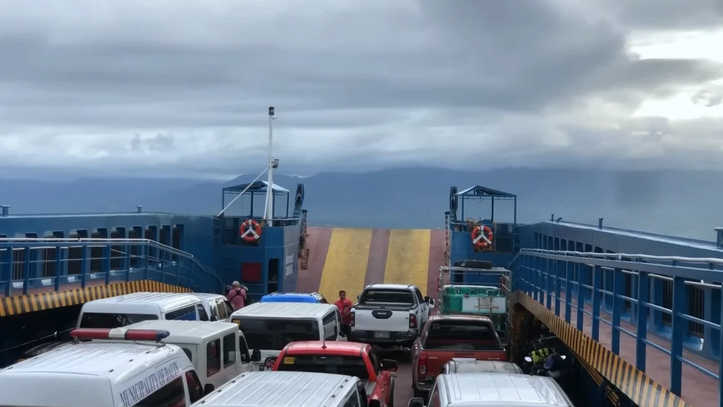 POV shot from the deck of a blue Maayo Shipping Ro-Ro ferry loaded with cars and vans, looking out toward the mountains of Negros Oriental across the sea under a cloudy sky.