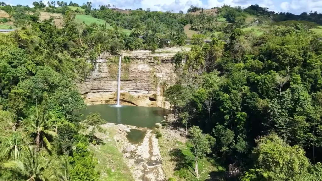 High-angle drone shot of Niludhan Falls in Bayawan City, showing a single thin stream of water cascading into a green natural pool surrounded by dense tropical forest and rocky cliffs.