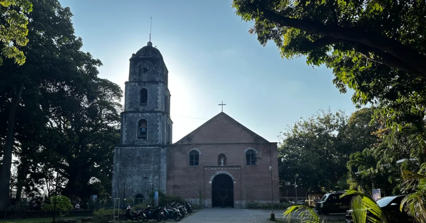 A historic stone and brick church in Bacong, Negros Oriental, with an old bell tower on the left, a cross above the main roof, motorcycles parked near the entrance, and large green trees framing the scene under a clear blue sky.