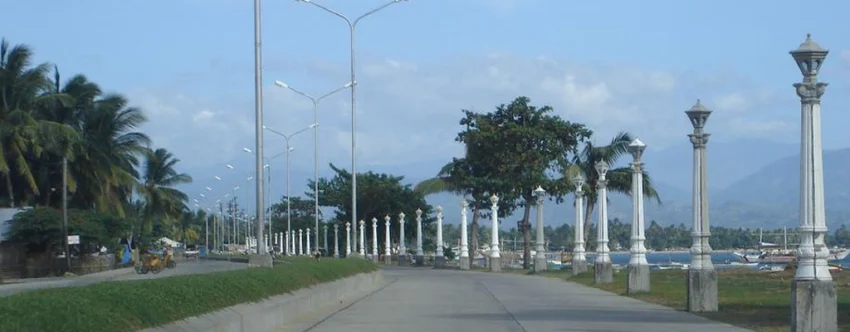 Bayawan Boulevard in Bayawan City, Negros Oriental, showing a wide seaside walkway lined with white lamp posts, palm trees, streetlights, nearby fishing boats, and distant mountain views under a blue sky.