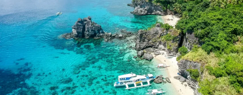 Aerial view of a white outrigger boat anchored beside a small tropical beach with clear turquoise water, rocky cliffs, coral reefs, and lush green coastal trees in Dauin, Negros Oriental.
