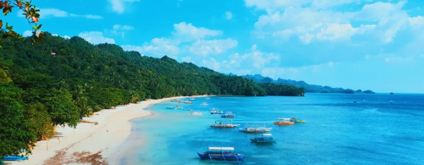 Wide coastal view of Sipalay City, Negros Occidental, showing a long white sand beach, bright blue water, several outrigger boats, green forested hills, and small islands under a clear tropical sky.
