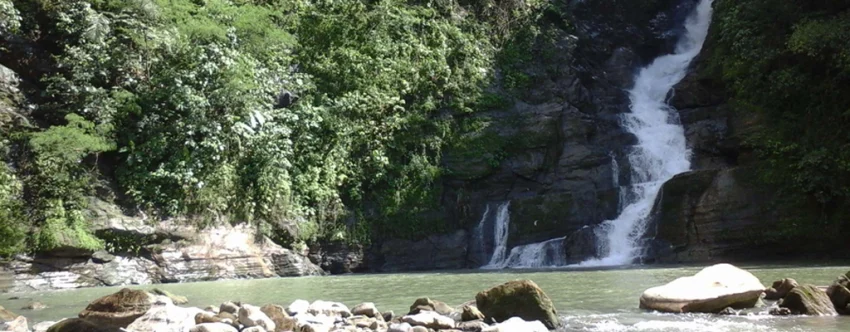 Talostos Falls in Sta. Catalina, Negros Oriental, with white water cascading down dark rock formations into a calm natural pool surrounded by large stones and dense green forest.