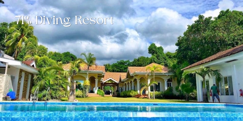 Featured image of 71% Diving Resort showing a bright blue swimming pool in front of tropical gardens, palm trees, and low-rise resort buildings with tiled roofs beneath a partly cloudy sky.