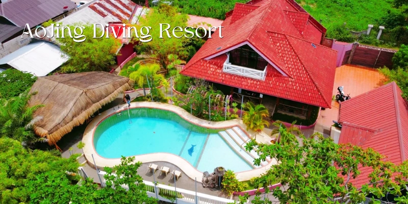 Featured image of AoJing Diving Resort showing an aerial view of a curved turquoise swimming pool surrounded by tropical plants, a thatched-roof pavilion, and red-roofed resort buildings.
