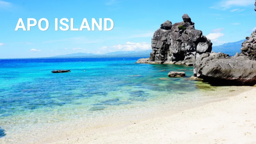 Featured image for an Apo Island Travel Guide showing a bright white-sand shoreline, clear turquoise and deep blue water, a small boat offshore, and large dark rock formations rising along the coast.