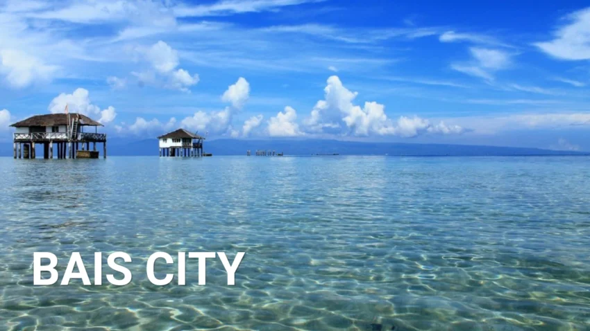 Bais City Travel Guide image showing clear shallow blue water, two nipa-style stilt cottages standing over the sea, distant mountains on the horizon, and a bright sky filled with soft white clouds.