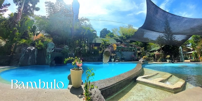 Featured image of Bambulo showing a large blue swimming pool surrounded by tropical plants, shaded lounging areas, decorative stone features, and a smaller shallow pool beneath a wide overhead sunshade.