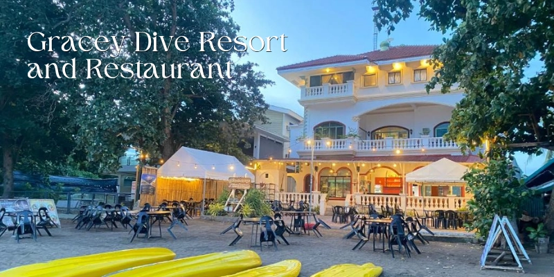 Featured image of Gracey Dive Resort and Restaurant showing a multi-story beachfront resort building with warm evening lights, outdoor dining tables on the sand, shaded seating areas, large trees, and yellow kayaks in the foreground.