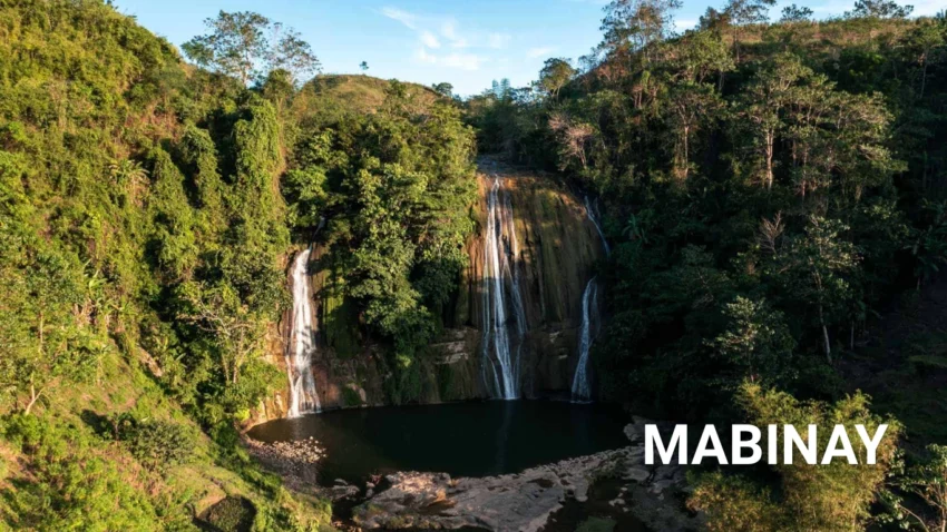 Featured image for a Mabinay Travel Guide showing a wide multi-stream waterfall cascading over a rocky cliff into a dark natural pool surrounded by dense green forest and rolling hills.