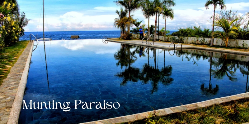 Featured image of Munting Paraiso showing a large oceanfront swimming pool with calm reflective water, palm trees along the edge, tropical landscaping, and an open view of the blue sea.