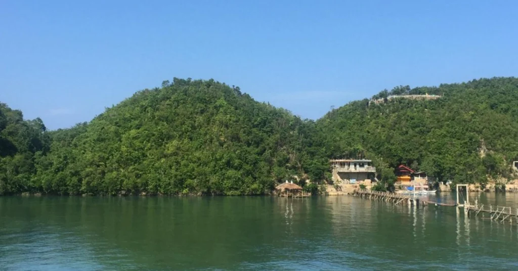 Tinagong Dagat Island Resort in Sipalay City beside a calm green lagoon, with forest-covered hills, waterfront buildings, a thatched cottage, and wooden walkways extending over the water.
