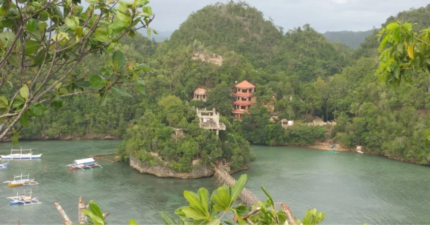 Tinagong Dagat Island Resort: Elevated view of a calm green lagoon surrounded by forested hills, with outrigger boats, a rocky islet, a wooden footbridge, and red-roofed resort buildings among the trees.